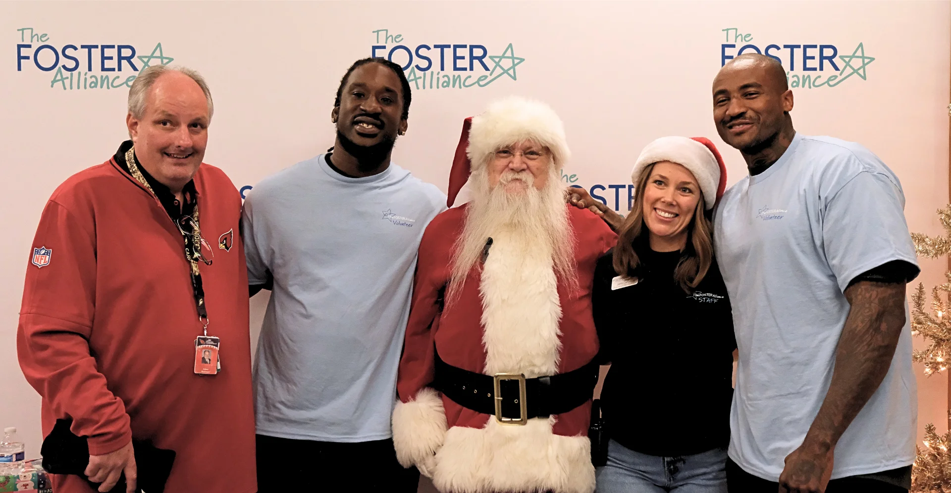 NFL player Markus Golden posing with Santa Claus and volunteers in front of The Foster Alliance backdrop during a holiday gift distribution event.